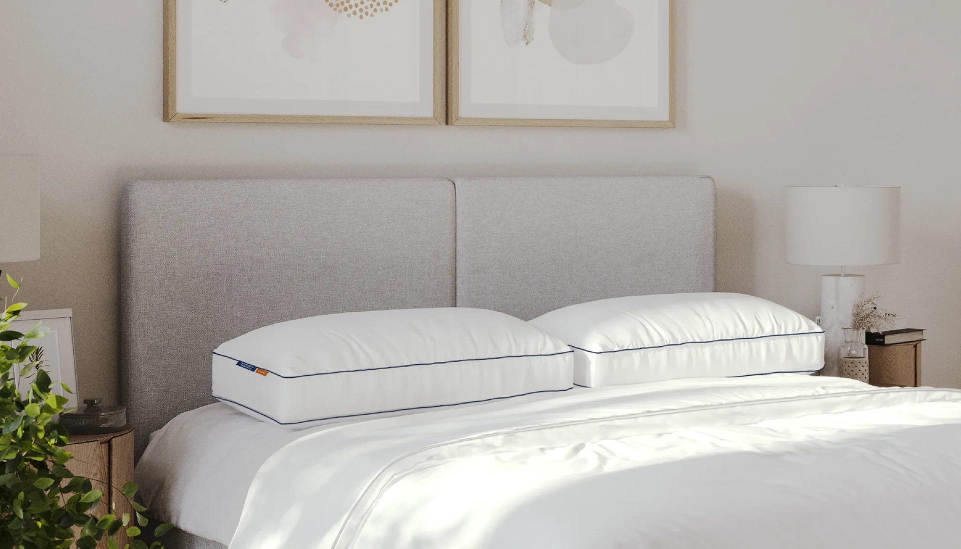 Twee Emma Premium Wolkenkussens v1 met witte hoes en blauwe bies, liggend naast elkaar op een opgemaakt bed met witte dekens en een lichtgrijze gestoffeerde hoofdsteun. De slaapkamer heeft een minimalistisch design met houten nachtkastjes, witte lampen en decoratieve elementen zoals een plant en abstracte kunst aan de muur.