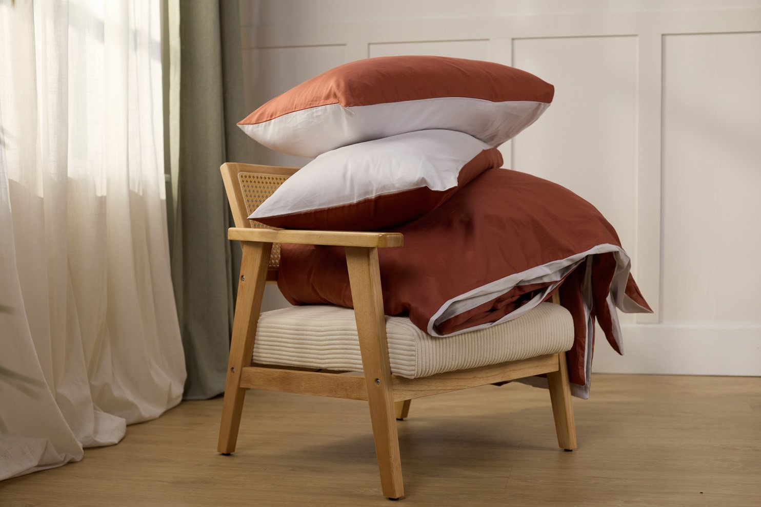 Stapel roestkleurig en wit beddengoed op een houten stoel met gecapitonneerde zitting, in een lichte slaapkamer setting.