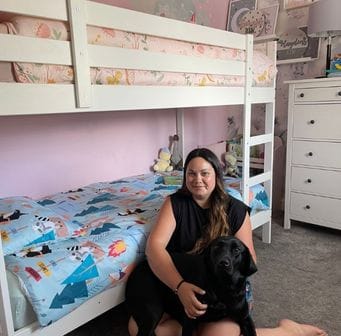 a white bunk bed with blue bedding on the lower bunk and pink bedding on the upper bunk, while a person sits on the floor in front of it holding a dog.