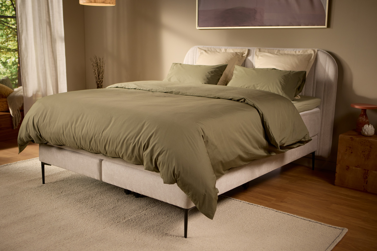 Schuin aanzicht van het lichtbeige gestoffeerde boxspringbed met kussens en beddengoed, in een zacht verlichte slaapkamer met warme tinten en natuurlijk ochtendlicht.