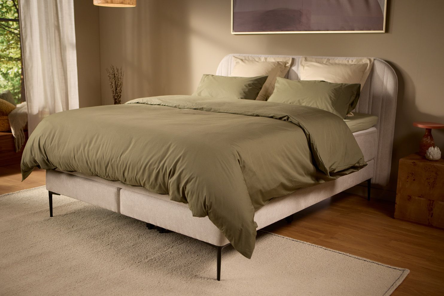 Schuin aanzicht van het lichtbeige gestoffeerde boxspringbed met kussens en beddengoed, in een zacht verlichte slaapkamer met warme tinten en natuurlijk ochtendlicht.