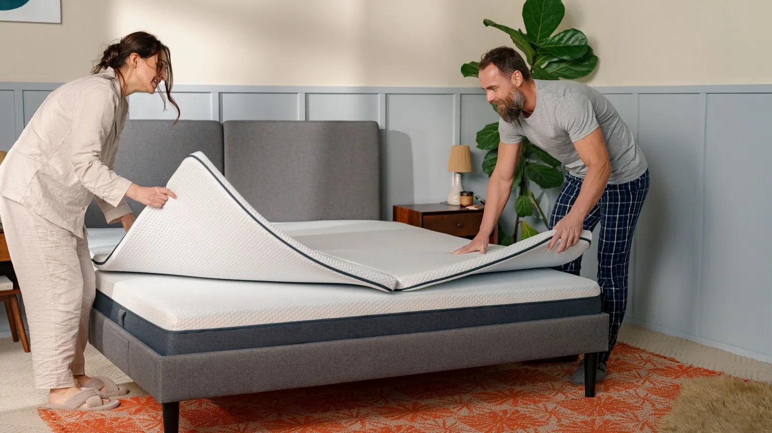Twee personen maken samen een bed op en leggen een Emma Flip Topper op een matras; het bed heeft een grijze stoffen hoofdsteun en bedframe, met een nachtkastje, lamp en plant rechts; de kamer is licht en sfeervol ingericht met een opvallend oranje-wit tapijt.