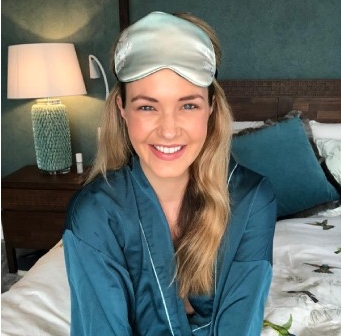 Woman sitting on a bed wearing a blue satin robe and a sleep mask, with a teal pillow on the bedroom setting.