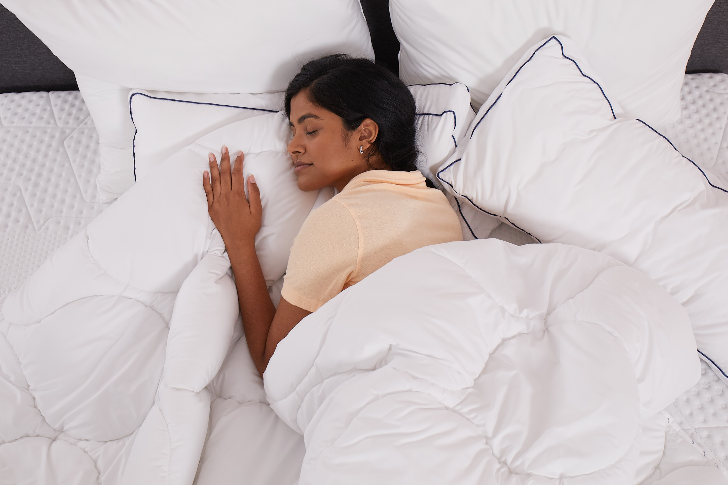 Persona sdraiata a letto sotto un piumone bianco, circondata da cuscini bianchi