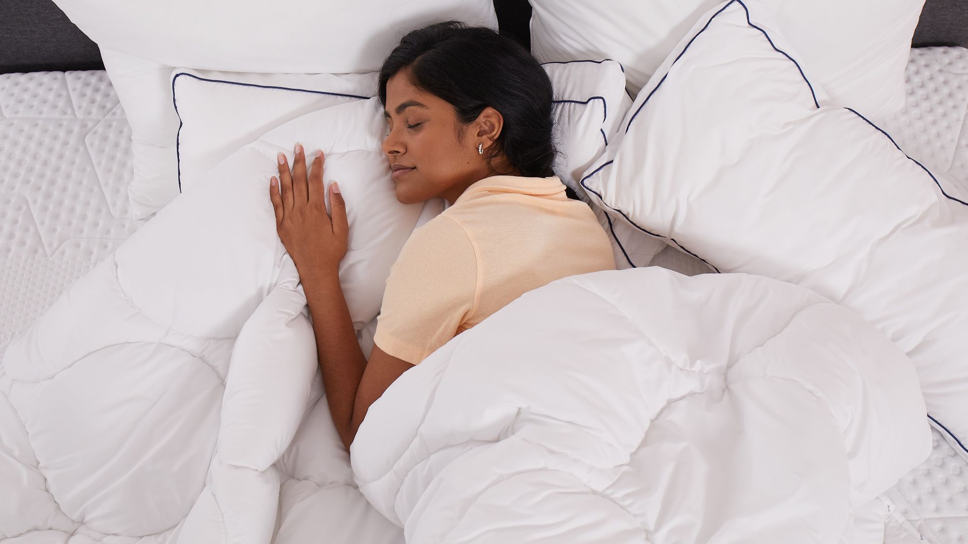 Persona sdraiata a letto sotto un piumone bianco, circondata da cuscini bianchi