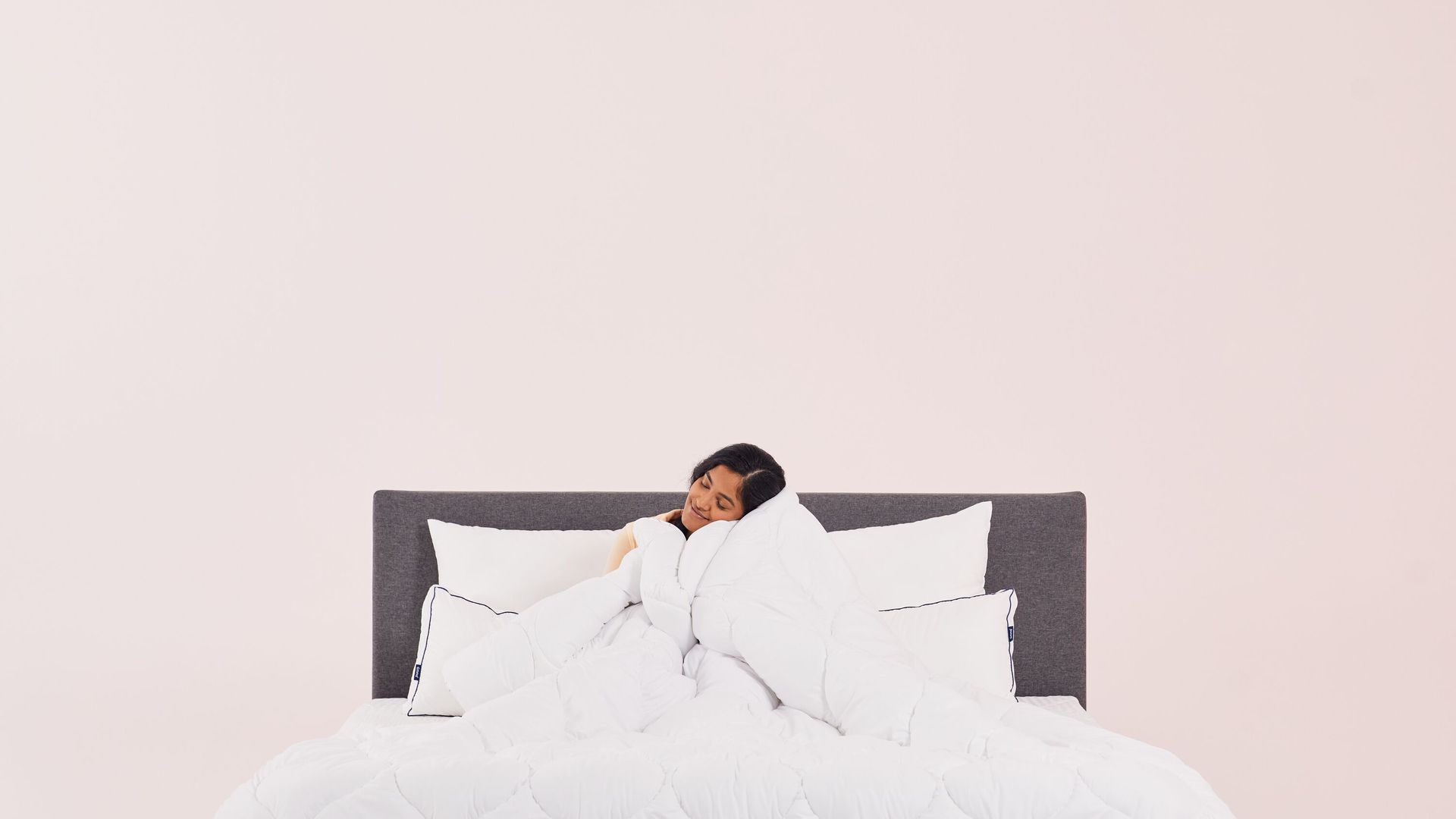 Persona seduta a letto con piumone bianco, testiera grigio scuro e sfondo chiaro.