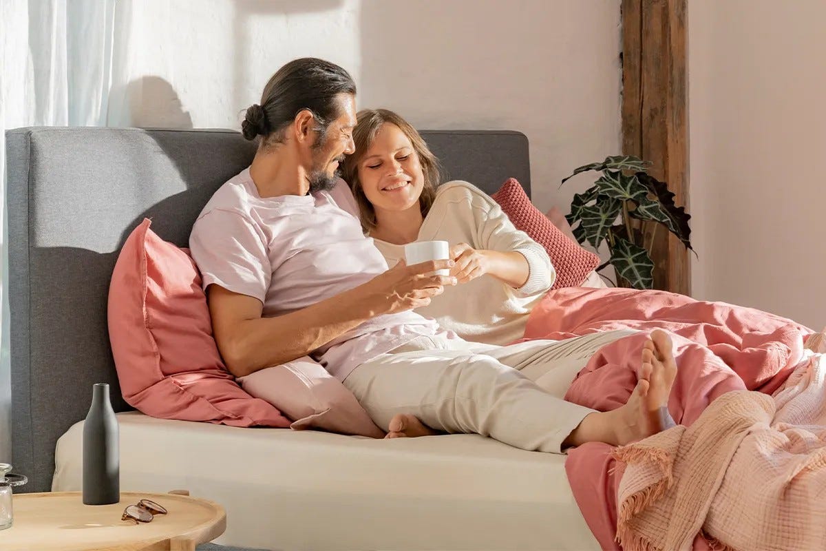 Due persone sedute su un letto con biancheria rosa e bianca, una tiene un libro.