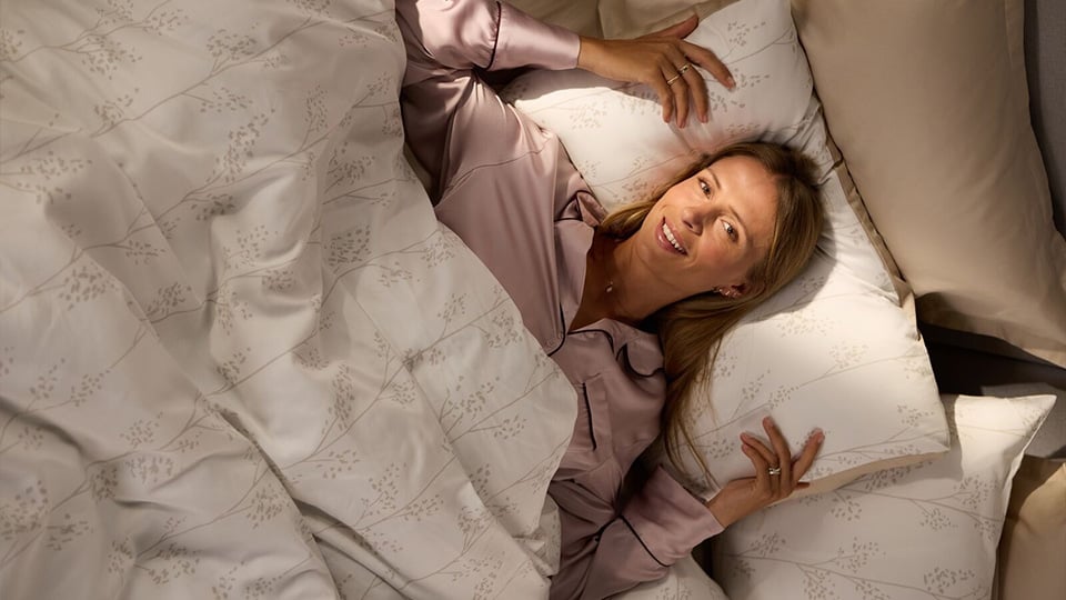 Woman lying under a beige floral duvet cover, holding a matching pillow.