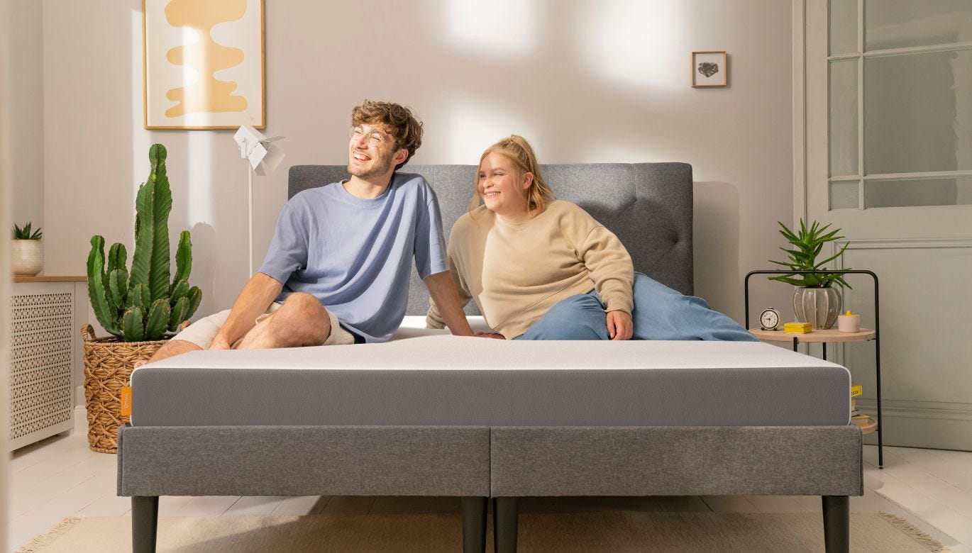 Twee personen zitten op bed met grijs hoofdbord en matrasbeschermer—slaapkamer met planten, wanddecoratie en nachtkastje met klok en kaars; natuurlijke lichtinval en rustige sfeer.