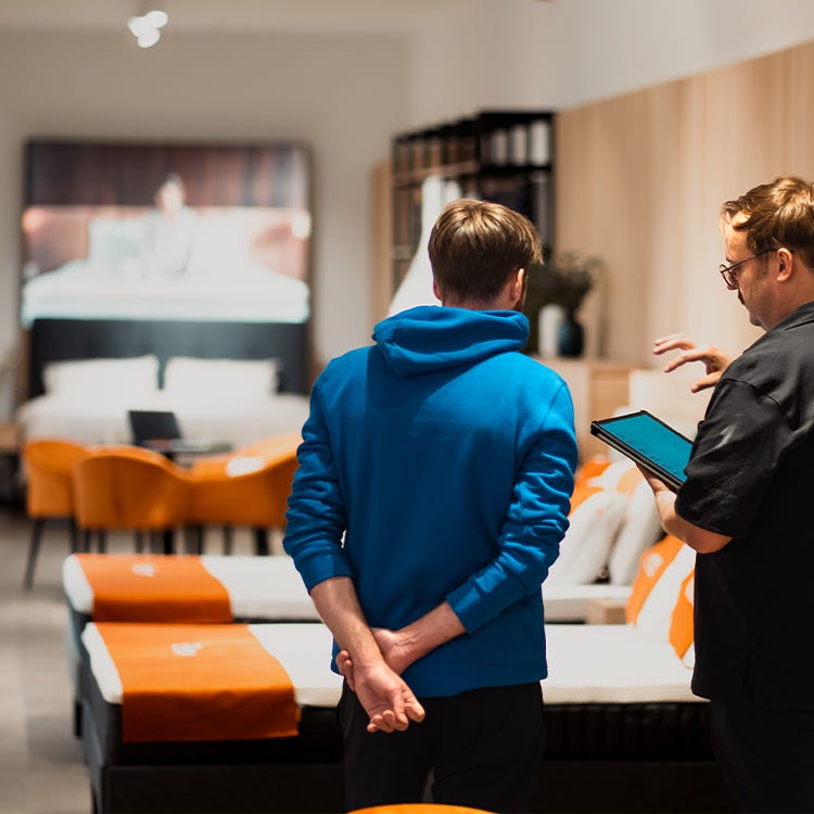 Twee winkelmedewerkers bespreken een matrasselectie; één toont informatie op een tablet terwijl de ander aandachtig luistert. Achter hen staan matrassen opgesteld en oranje stoelen bieden een comfortabele winkelomgeving.