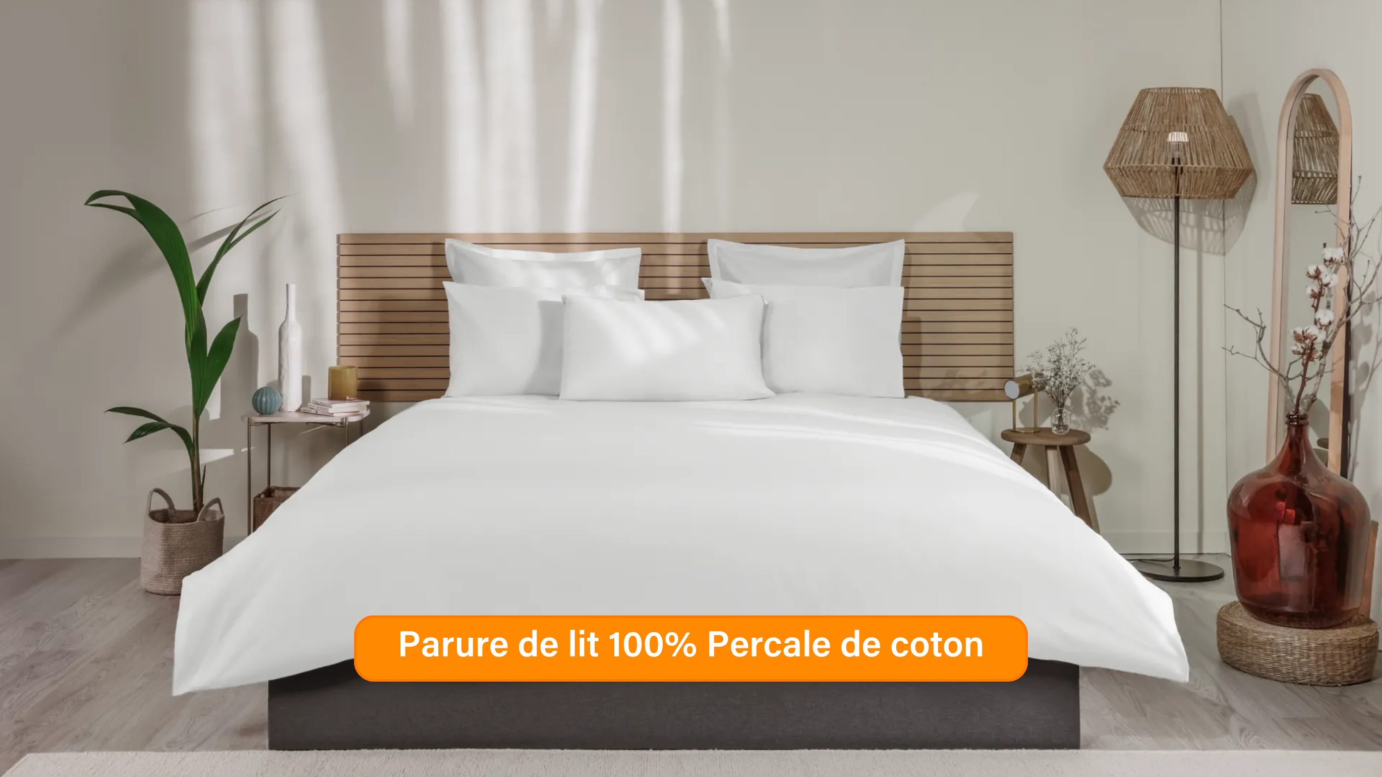 Chambre élégante avec lit soigneusement préparé, habillé d’une parure de lit 100 % satin de coton de couleur blanche ou beige clair. Plusieurs oreillers sont disposés contre un mur à panneaux. De chaque côté du lit, des tables de chevet avec lampe et objets décoratifs. Un banc rose est placé au pied du lit, assorti à un tabouret près de la table de chevet. Le sol en bois clair et les murs neutres créent une ambiance douce et raffinée.