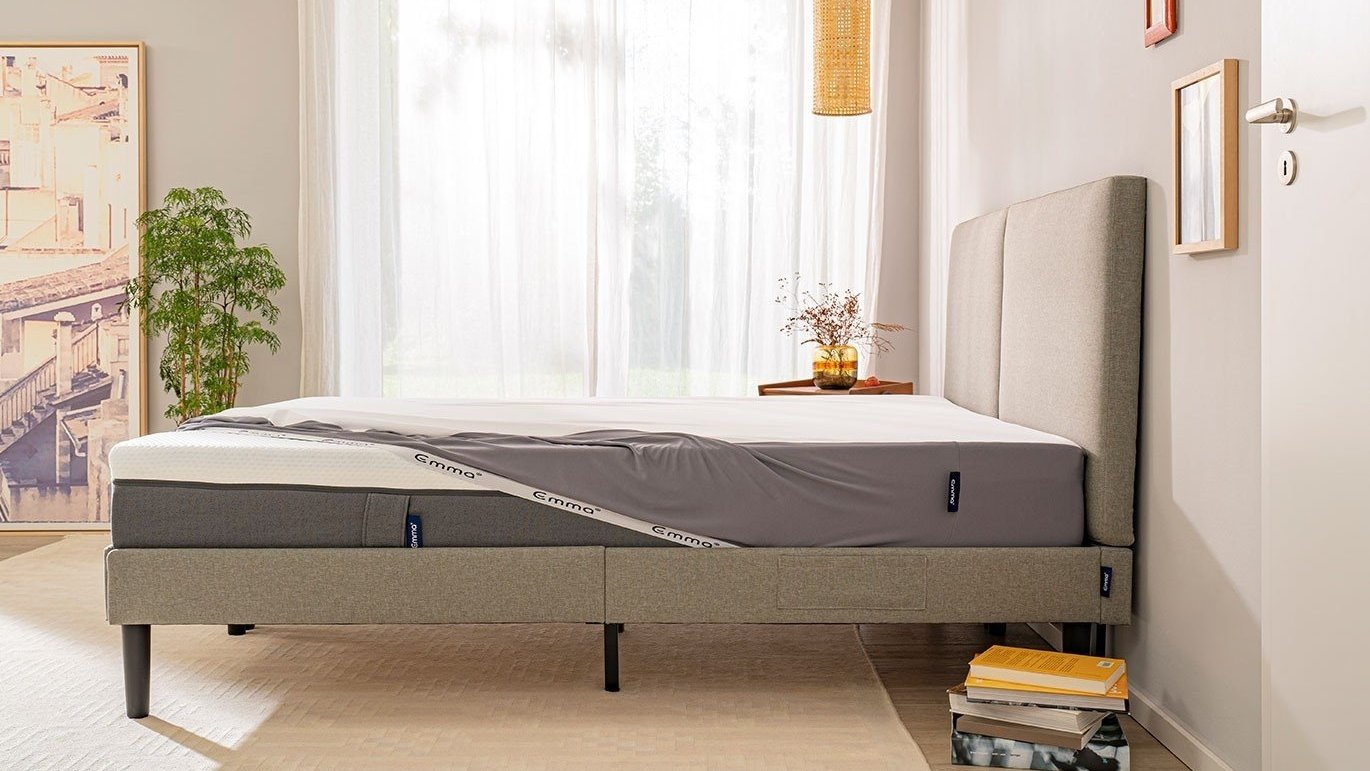 Moderne slaapkamer met grijs gestoffeerd bedframe en hoofdbord; Emma-matras herkenbaar aan merklabels; kamer wordt verlicht door daglicht via een groot raam met witte vitrage; linkerzijde toont ingelijst stadszicht en groene kamerplant; rechterzijde heeft houten nachtkastje met vaas en gedroogde bloemen, en boeken gestapeld op beige vloerbedekking; neutraal kleurpalet met lichte grijstinten.