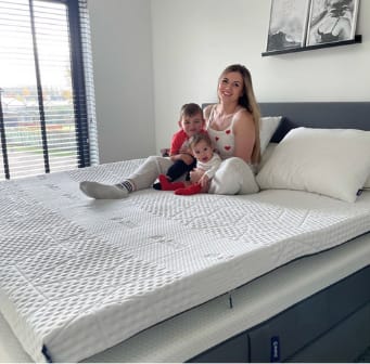 Moeder en twee kinderen zitten ontspannen op een bed met de Emma Original Pro Topper - een luxe topper ontworpen voor temperatuurregulatie en ergonomische ondersteuning tijdens gezinsmomenten.