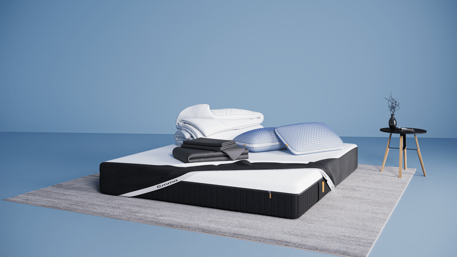 Ensemble de literie présenté dans une chambre moderne : matelas Hybride II ou Hybride Premium II avec base noire et surface blanche, partiellement recouvert d’un plaid gris foncé. Sur le matelas, deux oreillers Nuage Elite 40x70 cm bleus, une couette Nuage Tempérée blanche pliée, un protège-matelas, et une parure de lit en satin de coton blanc comprenant une housse de couette et des taies d’oreillers. Le tout est posé sur un tapis clair, avec une table d’appoint noire à pieds en bois et une décoration minimaliste.