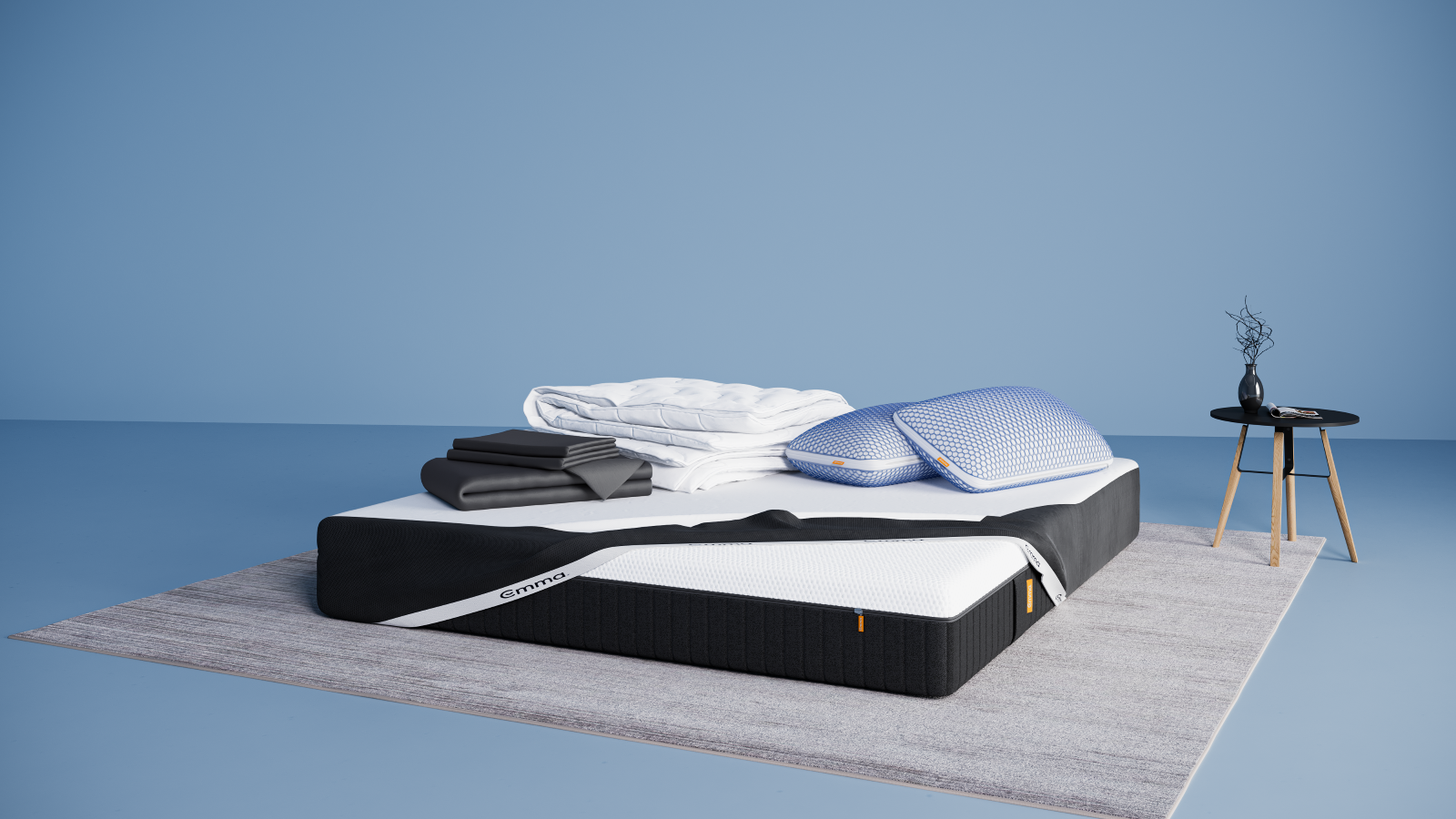 Ensemble de literie présenté dans une chambre moderne : matelas Hybride II ou Hybride Premium II avec base noire et surface blanche, partiellement recouvert d’un plaid gris foncé. Sur le matelas, deux oreillers Nuage Elite 40x70 cm bleus, une couette Nuage Tempérée blanche pliée, un protège-matelas, et une parure de lit en satin de coton blanc comprenant une housse de couette et des taies d’oreillers. Le tout est posé sur un tapis clair, avec une table d’appoint noire à pieds en bois et une décoration minimaliste.