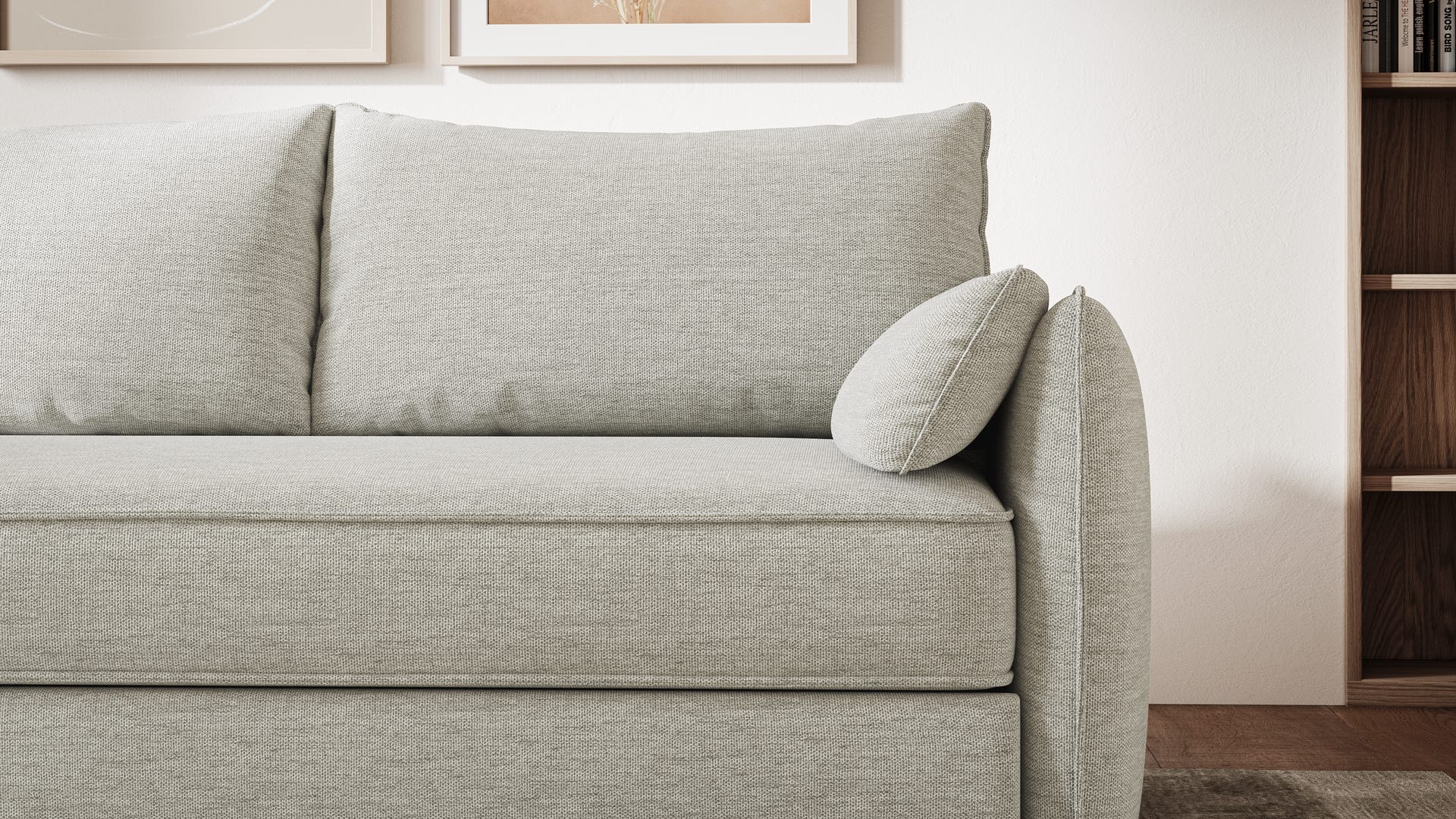 Vue rapprochée d’un canapé convertible gris clair au design épuré, avec dossier droit, assise unique et deux coussins de dossier. Un coussin d’accoudoir cylindrique est positionné verticalement. En arrière-plan, mur avec cadre décoratif et étagère en bois avec objets de décoration. Ambiance moderne et minimaliste.