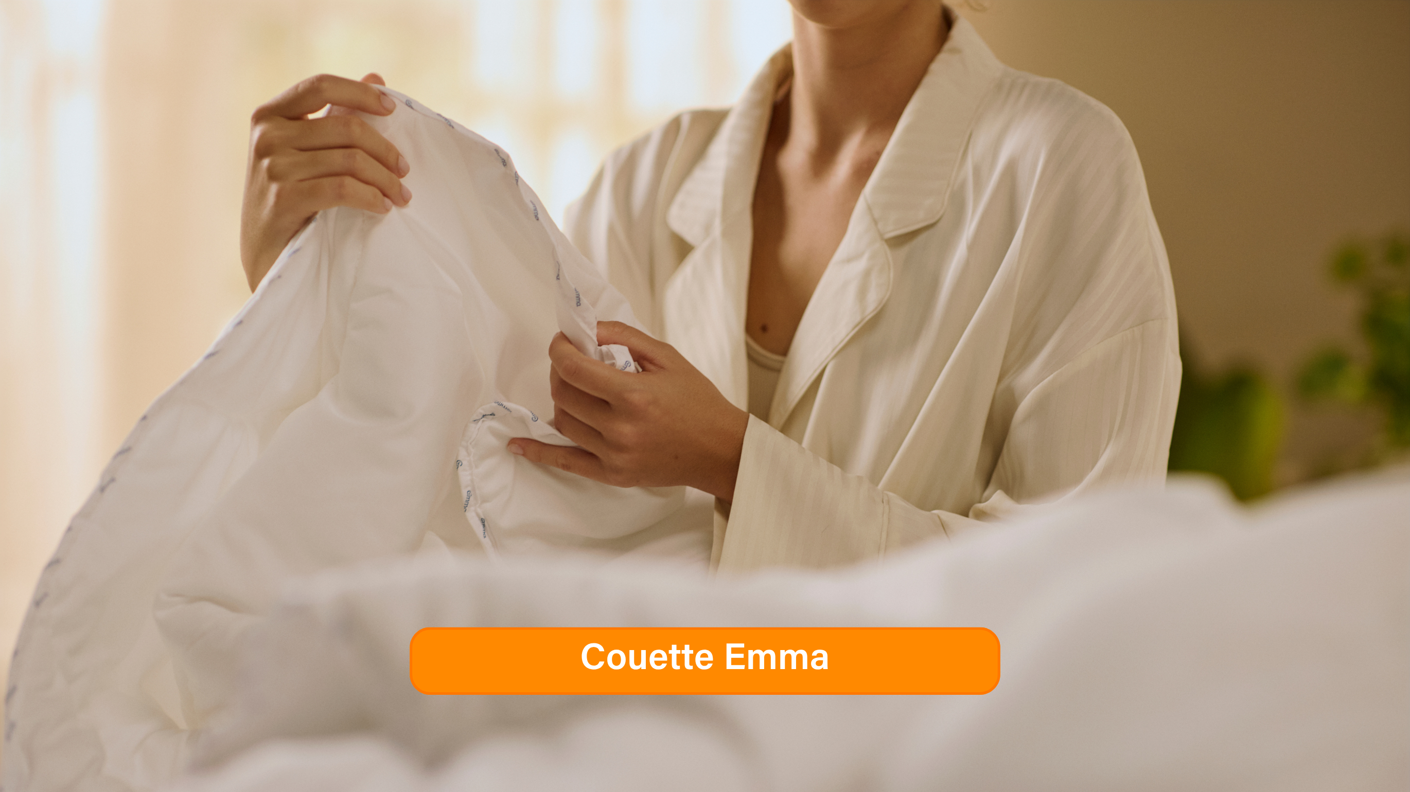 Gros plan sur la Couette Nuage Tempérée Emma, blanche, douce et moelleuse, avec une étiquette "Emma" cousue sur la couture. La texture lisse et les plis légers sont mis en valeur par un éclairage doux. Une bannière orange en bas de l’image indique "Couette Nuage