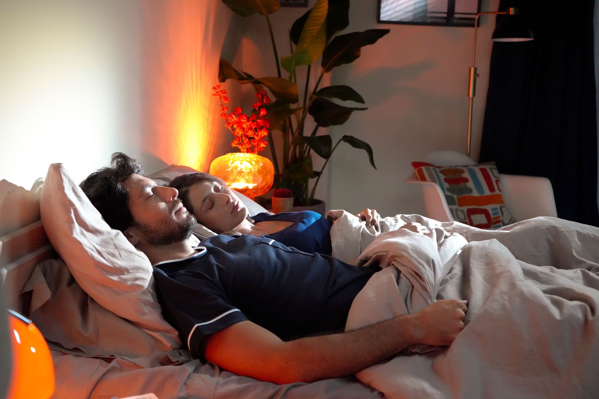 Echtpaar ligt op bed onder de deken van Emma Hug in warme slaapkamer met oranje lamp, planten en decoratieve accenten op achtergrond.