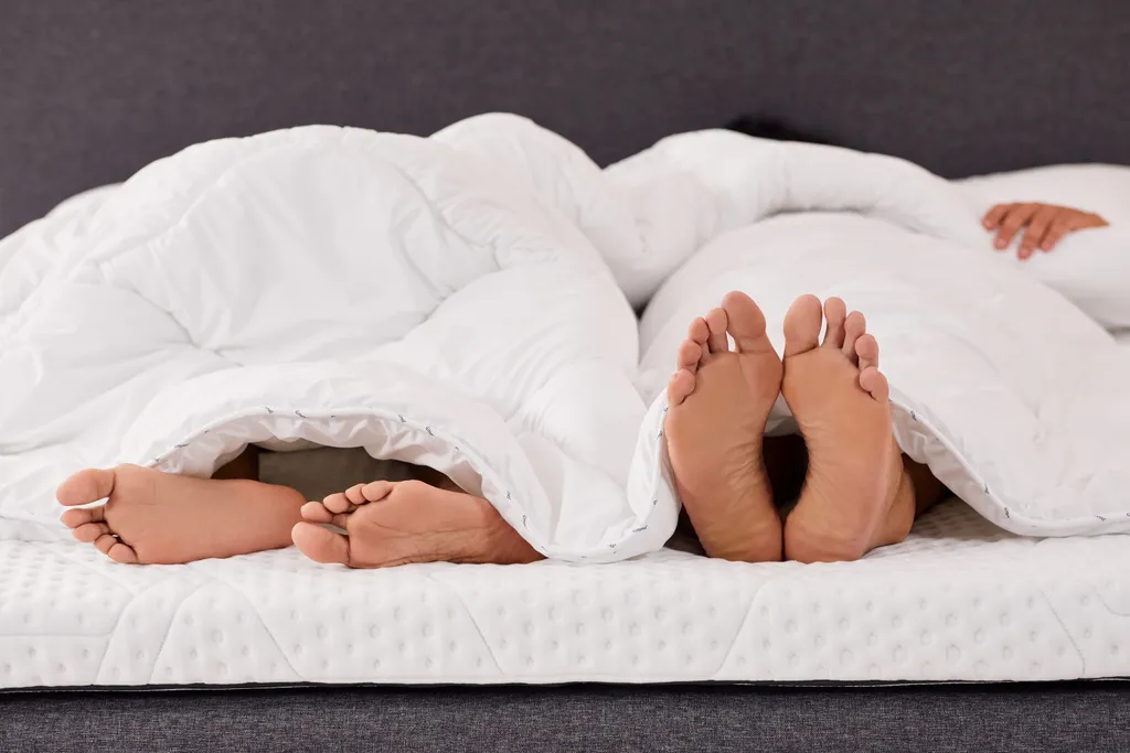 two pairs of feet peeking out from under a white comforter on a bed, with one hand resting on top of the blanket.