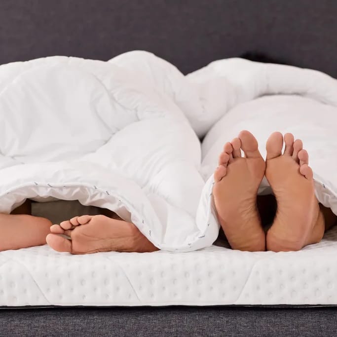 two pairs of feet peeking out from under a white comforter on a bed, with one hand resting on top of the blanket.