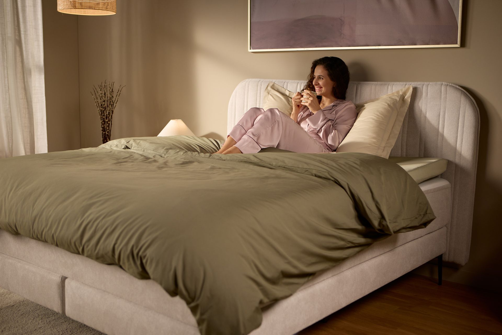 Schrägansicht des Bettes mit einer Person, die aufrecht am Kopfteil sitzt; zeigt Höhe und Halt bei weichem Morgenlicht.