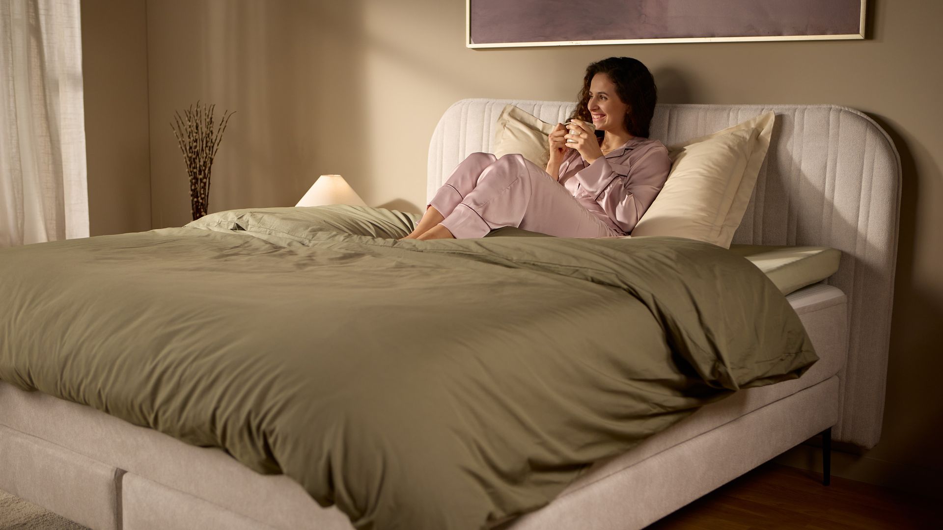 Schrägansicht des Bettes mit einer Person, die aufrecht am Kopfteil sitzt; zeigt Höhe und Halt bei weichem Morgenlicht.