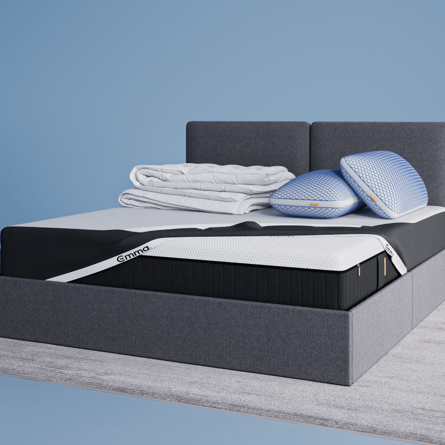 Un lit capitonné gris moderne avec des tiroirs de rangement, surmonté d'une literie et de coussins blancs, est posé sur un tapis blanc dans une chambre minimaliste aux murs bleu clair. Un petit tabouret avec une plante est placé à côté du lit.