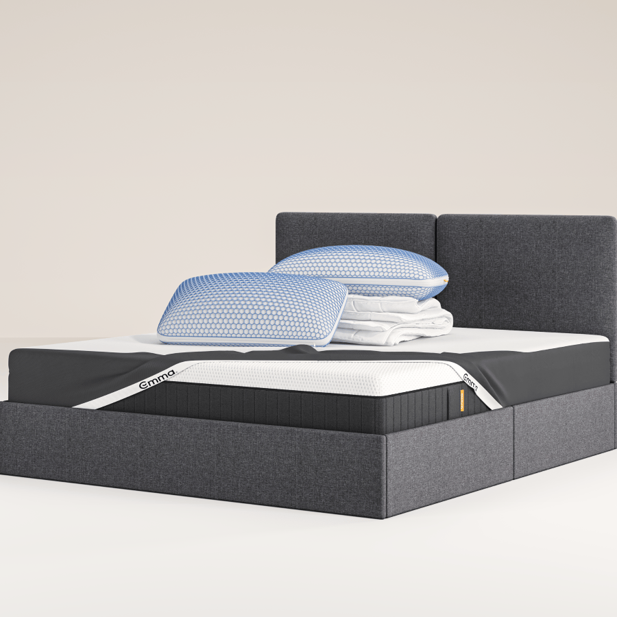 Un lit capitonné gris moderne avec des tiroirs de rangement, surmonté d'une literie et de coussins blancs, est posé sur un tapis blanc dans une chambre minimaliste aux murs bleu clair. Un petit tabouret avec une plante est placé à côté du lit.
