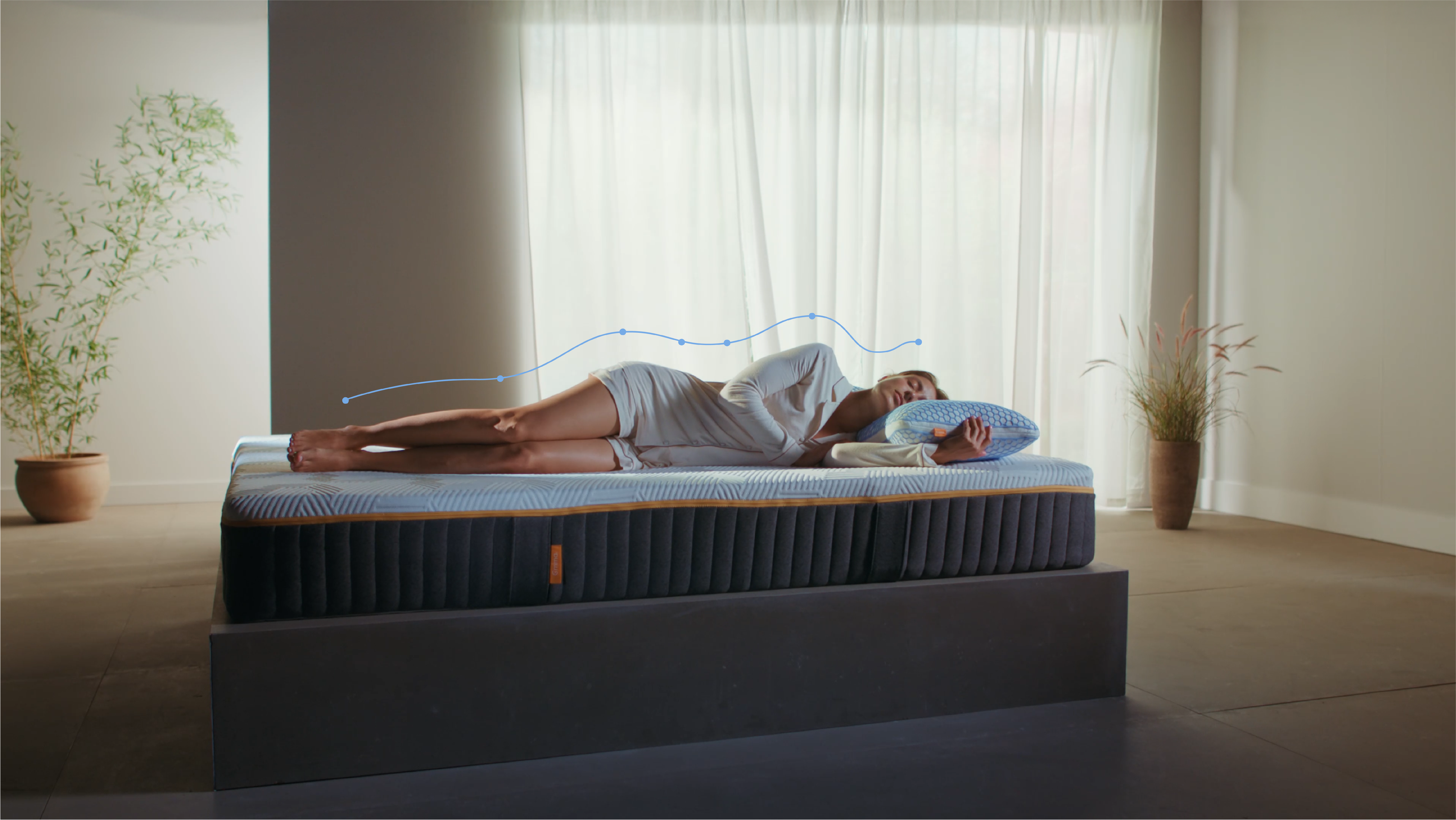 Persoon ligt op de zij op een matras in een minimalistische kamer; blauwe lijn met stippen volgt de contouren van het lichaam van hoofd tot voeten, wat de natuurlijke uitlijning van de ruggengraat benadrukt; omgeving bevat planten, lichtgordijnen en een platformbed.