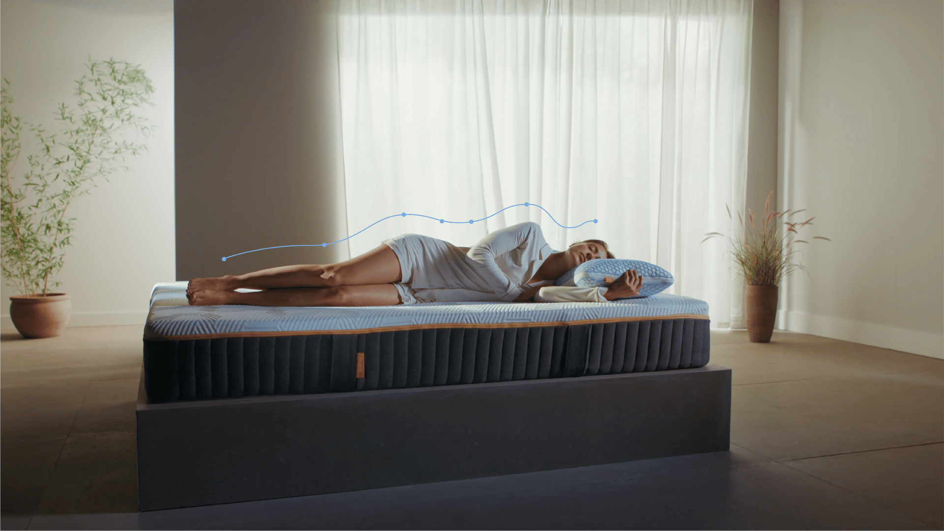 Persoon ligt op de zij op een matras in een minimalistische kamer; blauwe lijn met stippen volgt de contouren van het lichaam van hoofd tot voeten, wat de natuurlijke uitlijning van de ruggengraat benadrukt; omgeving bevat planten, lichtgordijnen en een platformbed.