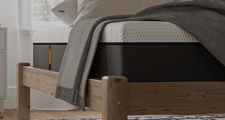Cama moderna com colchão alto Emma e cobertor cinza, ao lado de uma mesa de cabeceira clara com itens decorativos.