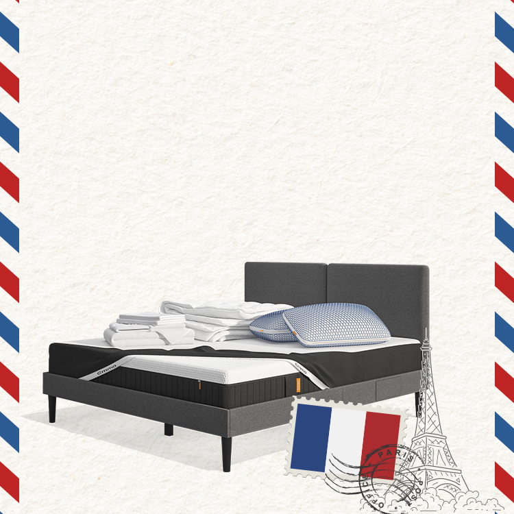 Lit moderne avec tête de lit grise et pieds noirs, recouvert de draps blancs. Deux oreillers blancs et une pile de linge plié sont posés sur le matelas. Arrière-plan beige avec une forme orange abstraite.