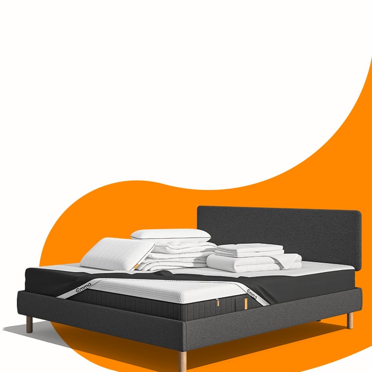 Lit moderne avec tête de lit grise et pieds noirs, recouvert de draps blancs. Deux oreillers blancs et une pile de linge plié sont posés sur le matelas. Arrière-plan beige avec une forme orange abstraite.