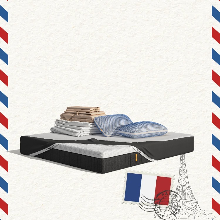 Lit moderne avec tête de lit grise et pieds noirs, recouvert de draps blancs. Deux oreillers blancs et une pile de linge plié sont posés sur le matelas. Arrière-plan beige avec une forme orange abstraite.