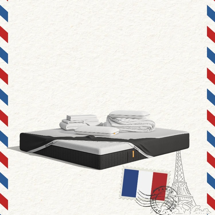 Lit moderne avec tête de lit grise et pieds noirs, recouvert de draps blancs. Deux oreillers blancs et une pile de linge plié sont posés sur le matelas. Arrière-plan beige avec une forme orange abstraite.