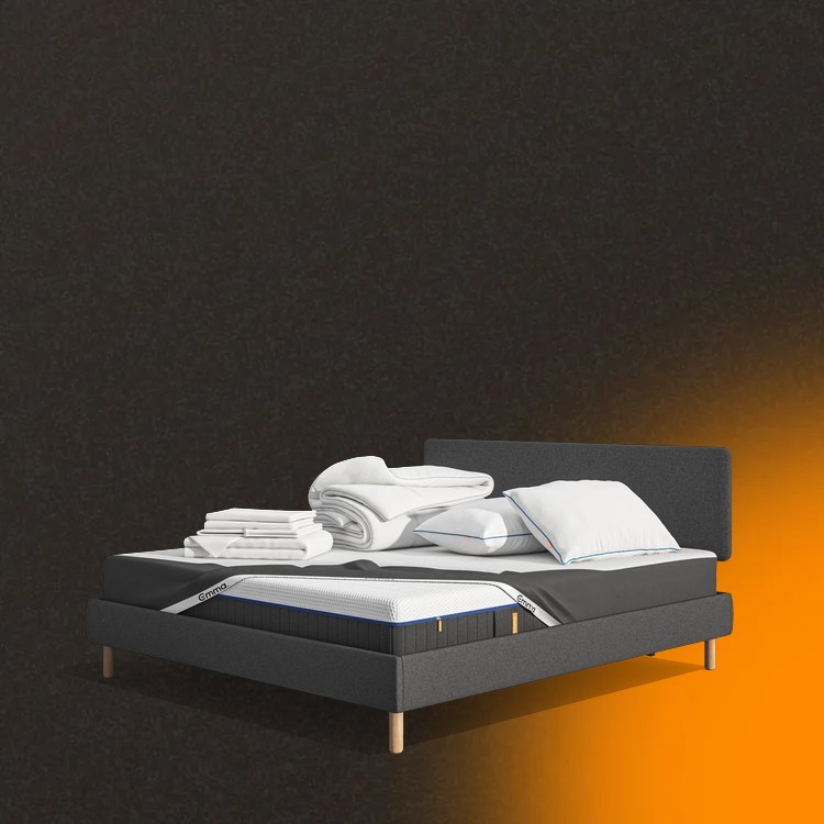Lit moderne avec tête de lit grise et pieds noirs, recouvert de draps blancs. Deux oreillers blancs et une pile de linge plié sont posés sur le matelas. Arrière-plan beige avec une forme orange abstraite.