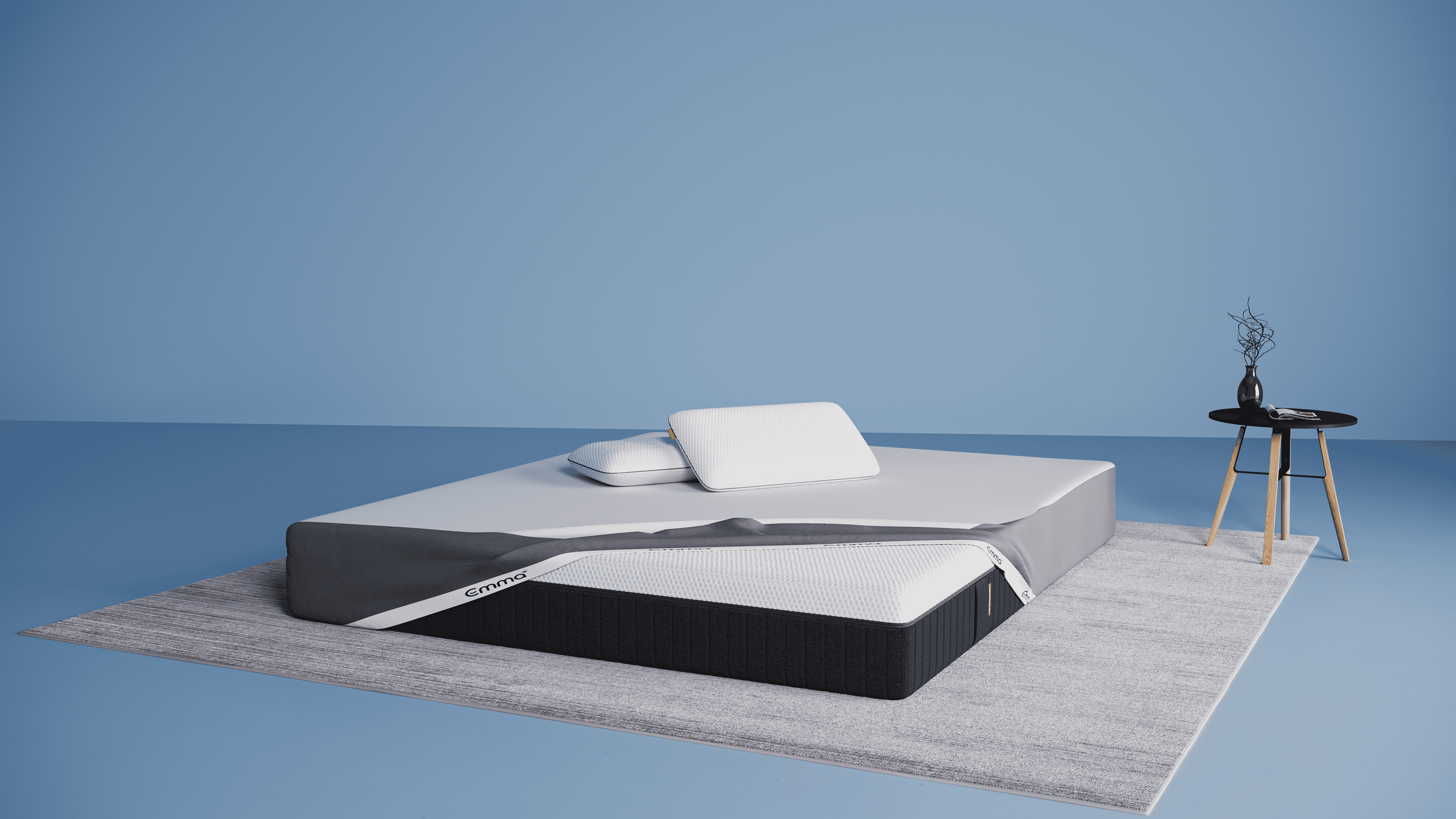 Minimalistische slaapkamer met een Emma Hybrid Premium matras op een lichtgrijs tapijt, deels bedekt met een grijze matrasbeschermer. Op het matras liggen twee witte Emma Diamond Kussens met zwarte rand. Rechts staat een zwart bijzettafeltje met houten poten, waarop een zwarte vaas met decoratieve takjes en een tijdschrift liggen. Deze afbeelding vertegenwoordigt de Emma Hybrid Premium Deal met gratis matrasbeschermer, Wolkenkussen(s) en Hybrid Premium matras.