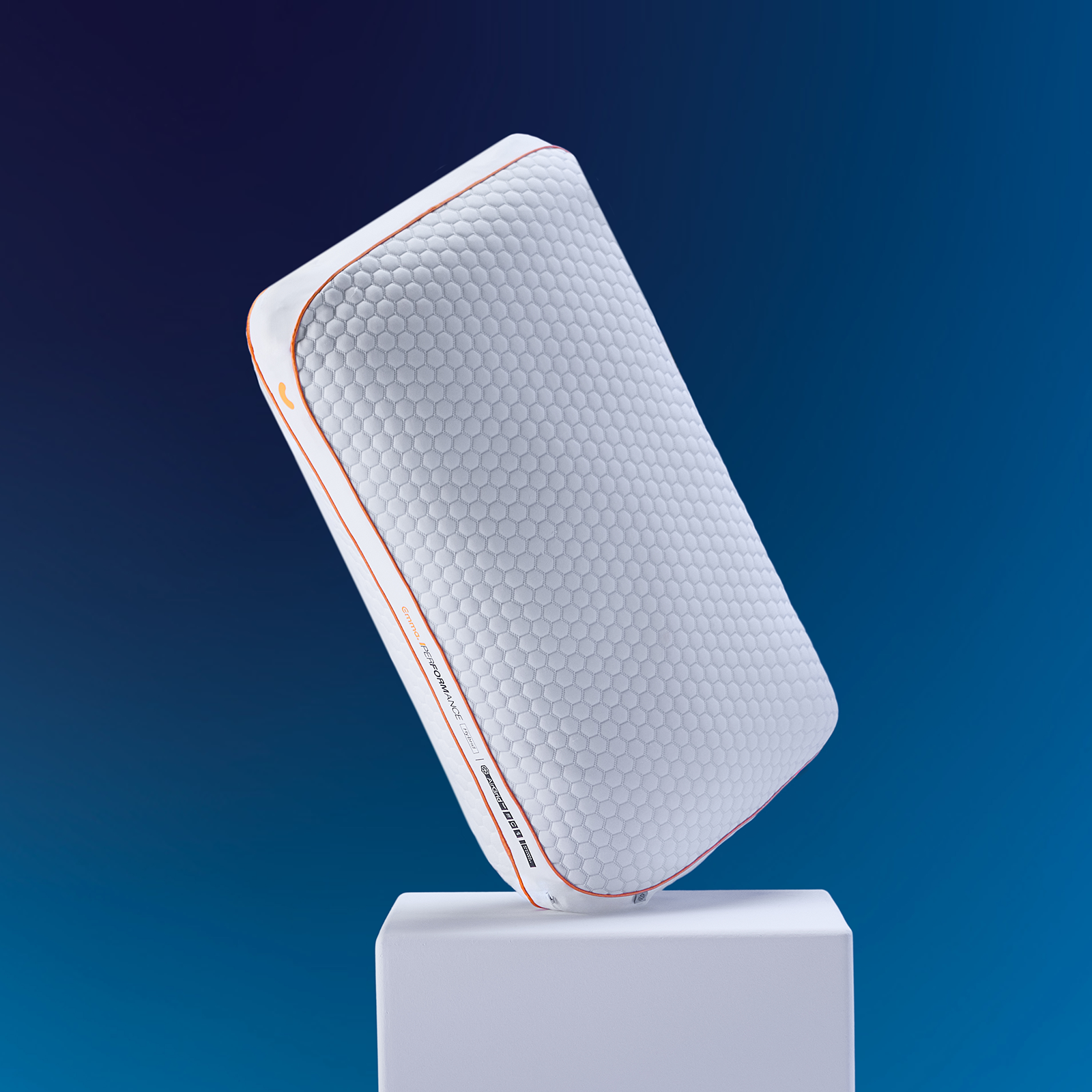 Emma Performance Hybrid Pillow tilted on a white plinth showing hexagonal cover and orange trim against a blue background.