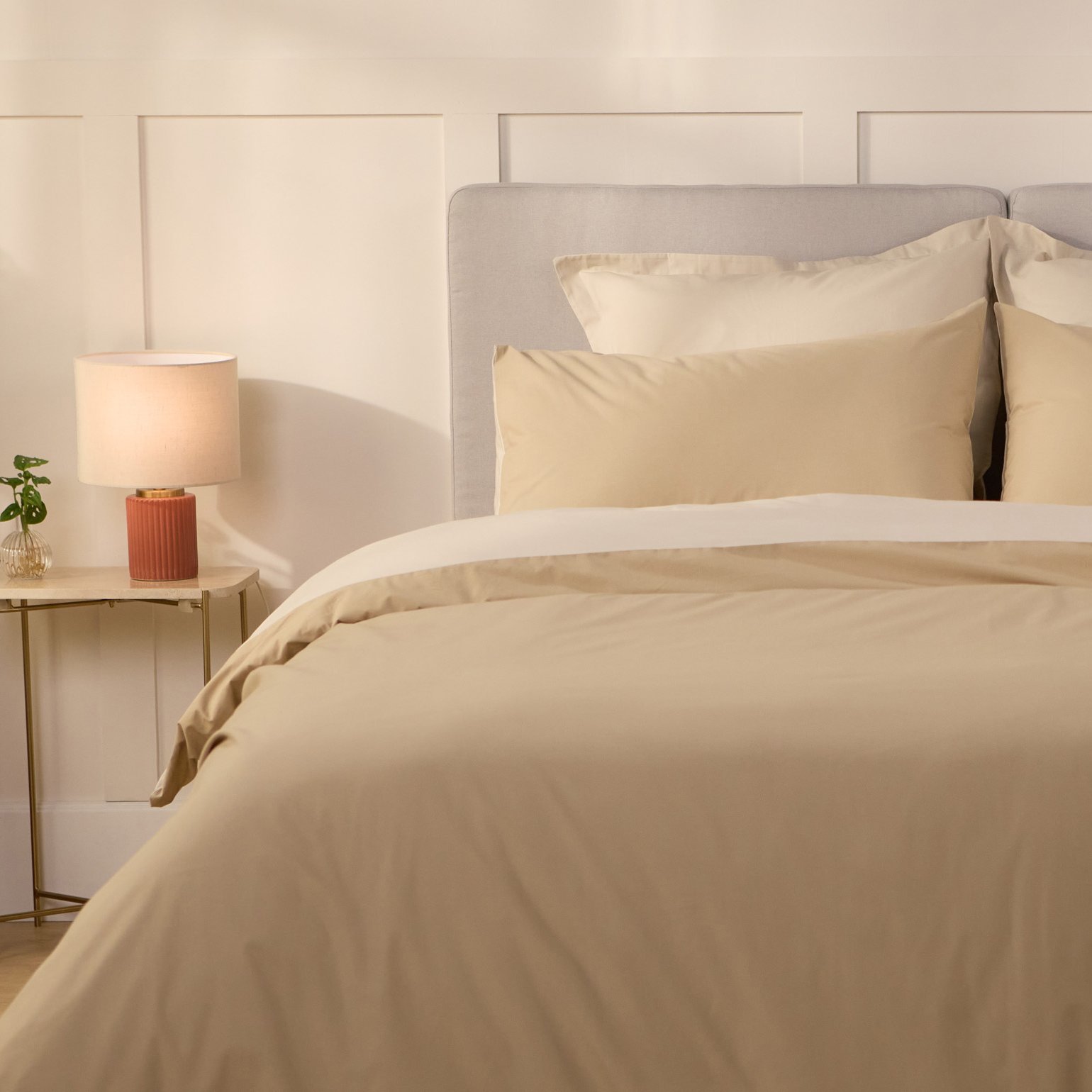 Opgemaakt bed met beige beddengoed en meerdere kussens, naast een bijzettafel met lamp en plant in een lichte, warme slaapkamer.