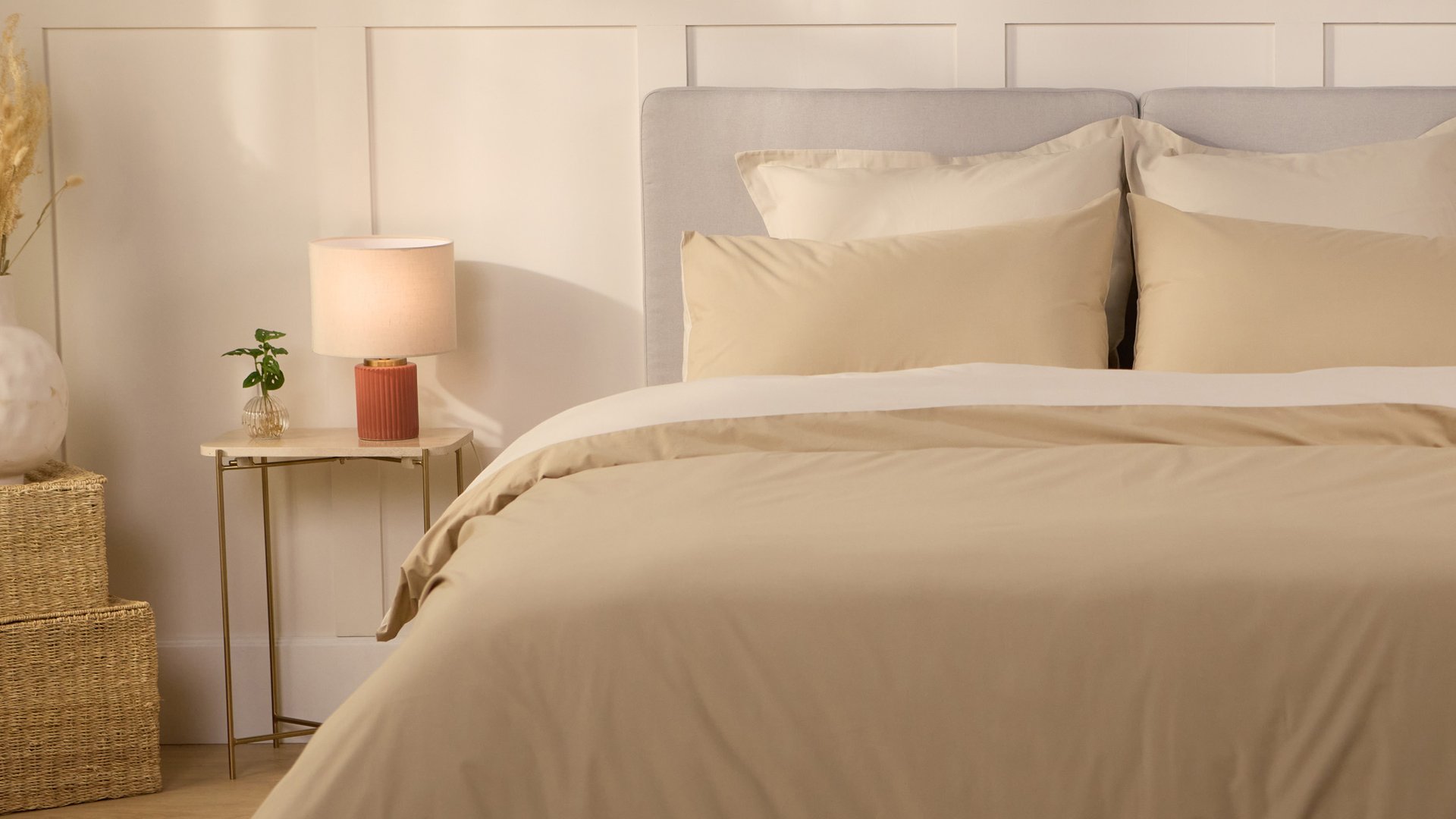 Opgemaakt bed met beige beddengoed en meerdere kussens, naast een bijzettafel met lamp en plant in een lichte, warme slaapkamer.
