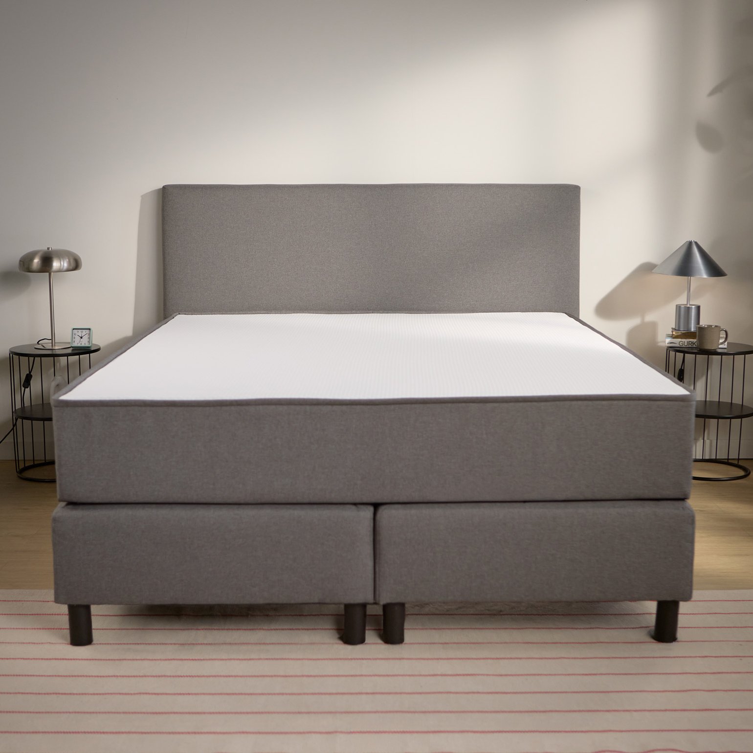 Vooraanzicht van het bed met gestoffeerde boxspringbasis, hoog hoofdbord en gelijkmatig geplaatste steunpoten.