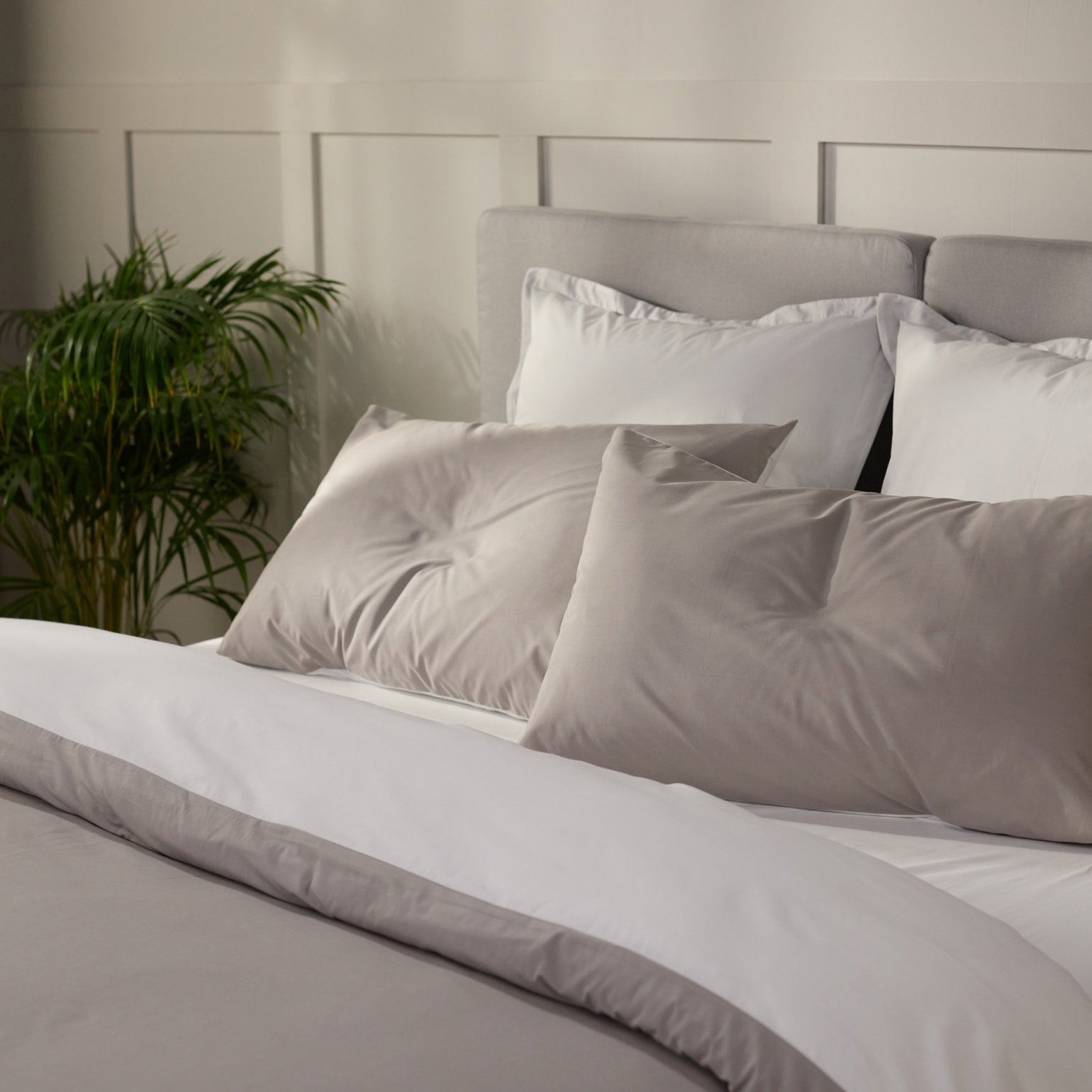 Opgemaakt bed met lichtgrijs en wit beddengoed en meerdere kussens, met een groene kamerplant en een houten stoel in een lichte slaapkamer.