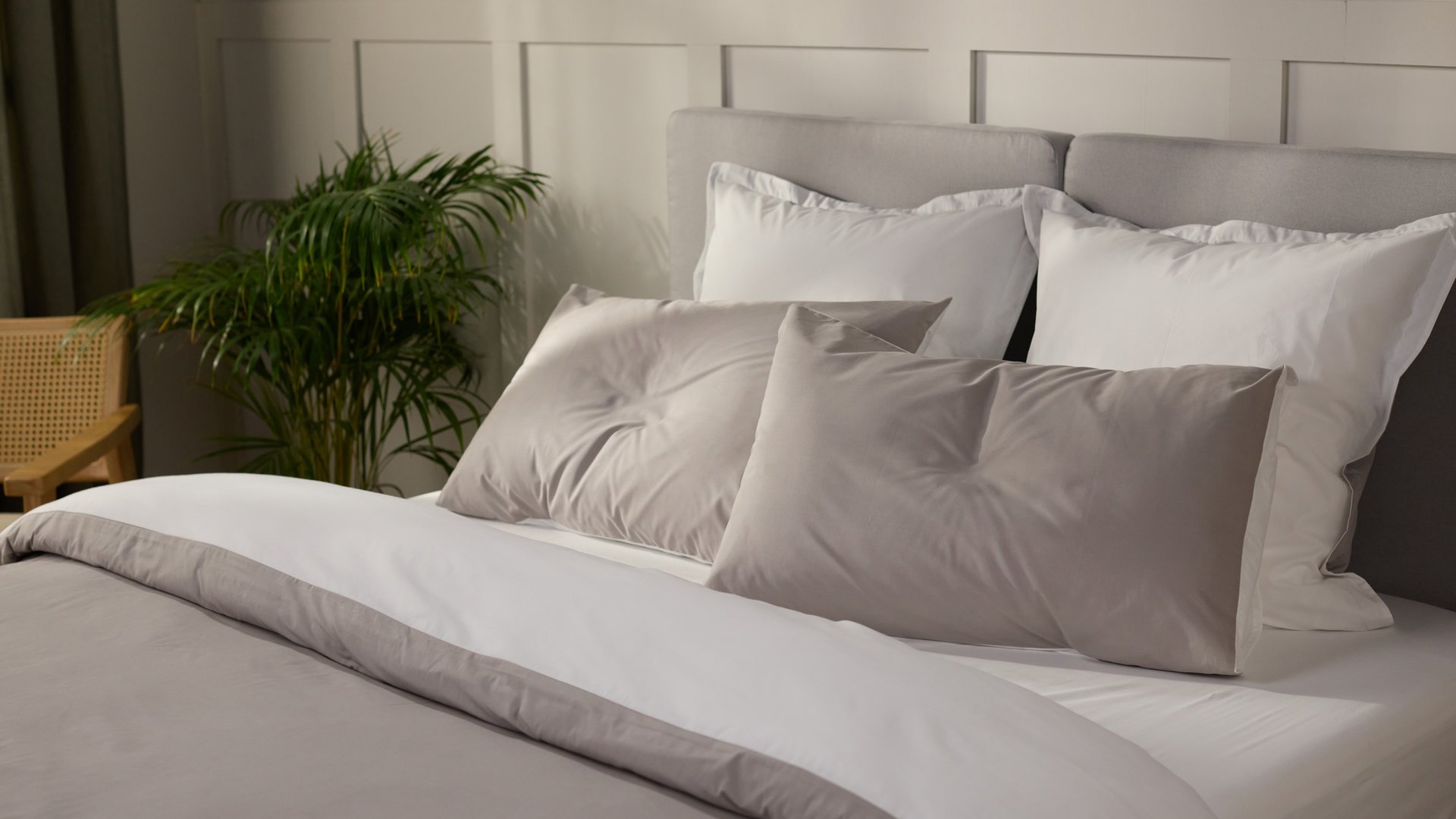 Opgemaakt bed met lichtgrijs en wit beddengoed en meerdere kussens, met een groene kamerplant en een houten stoel in een lichte slaapkamer.