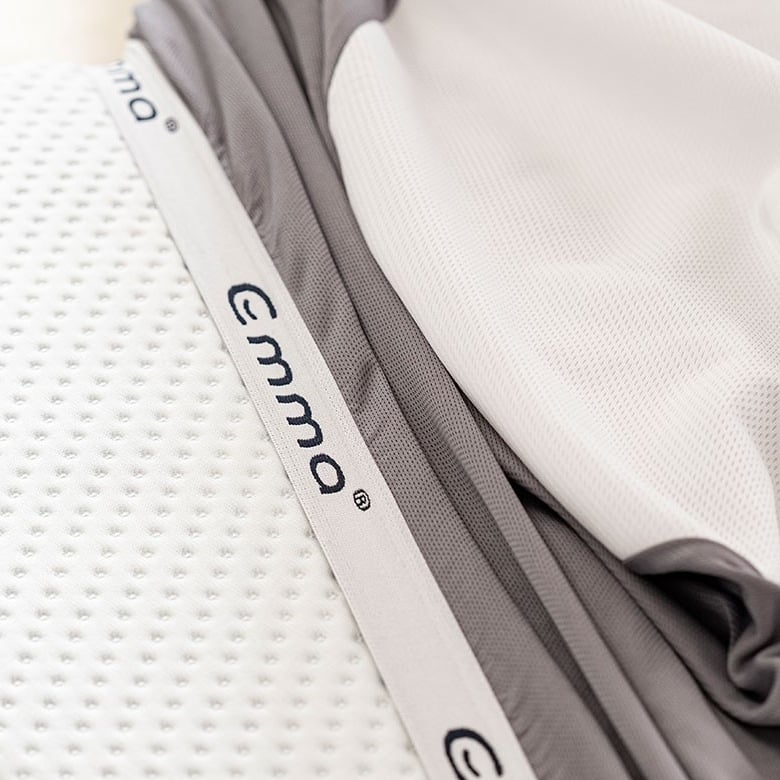 Close-up van een Emma-matras met een witte, gestructureerde bovenlaag en een grijs-witte rand; merknaam “Emma” zichtbaar in blauwe letters op de rand van de matras; compositie toont alleen het matras zonder omgeving of bedframe.
