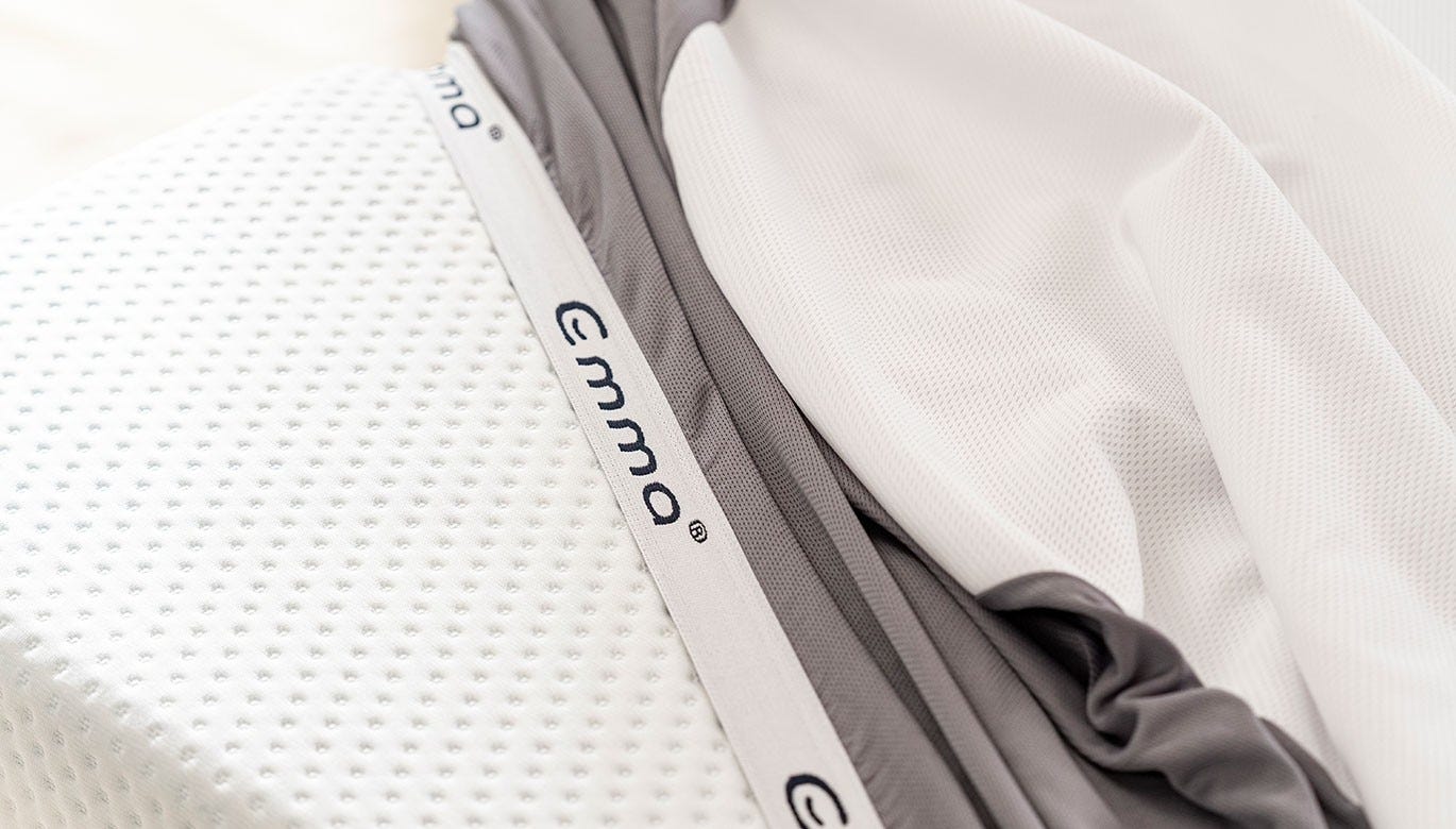 Close-up van de Emma Matrasbeschermer met wit geperforeerd oppervlak, grijze textielrand en zichtbaar Emma-label—ontworpen voor ademend, waterdicht en allergievrij slaapcomfort.