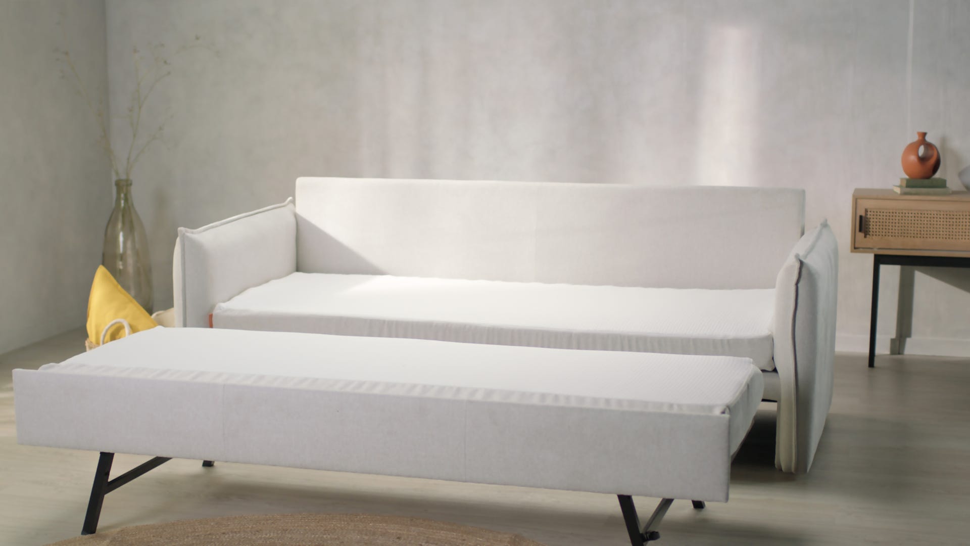 Canapé convertible en L, beige clair, déplié en position lit, avec pieds en métal noir. Placé sur un sol en bois clair avec tapis rond tressé partiellement visible. En arrière-plan, mur texturé, vase en verre avec branches séchées, coussin jaune au sol et console en bois avec vase et livre. Ambiance minimaliste, lumineuse et chaleureuse.