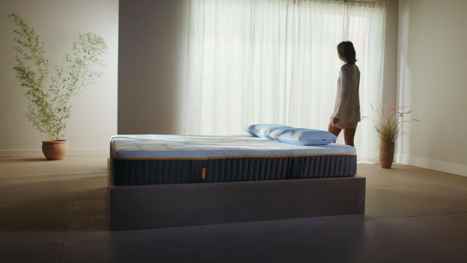Chambre moderne et épurée avec un lit équipé du Matelas Elite Zero Gravity II, composé d’un matelas bleu et blanc et de deux oreillers assortis. Le lit repose sur une base sombre. Une personne en tenue claire se tient près d’une grande fenêtre avec rideaux blancs, laissant entrer une lumière naturelle douce. Deux plantes en pot encadrent la pièce, créant une ambiance calme et sereine.