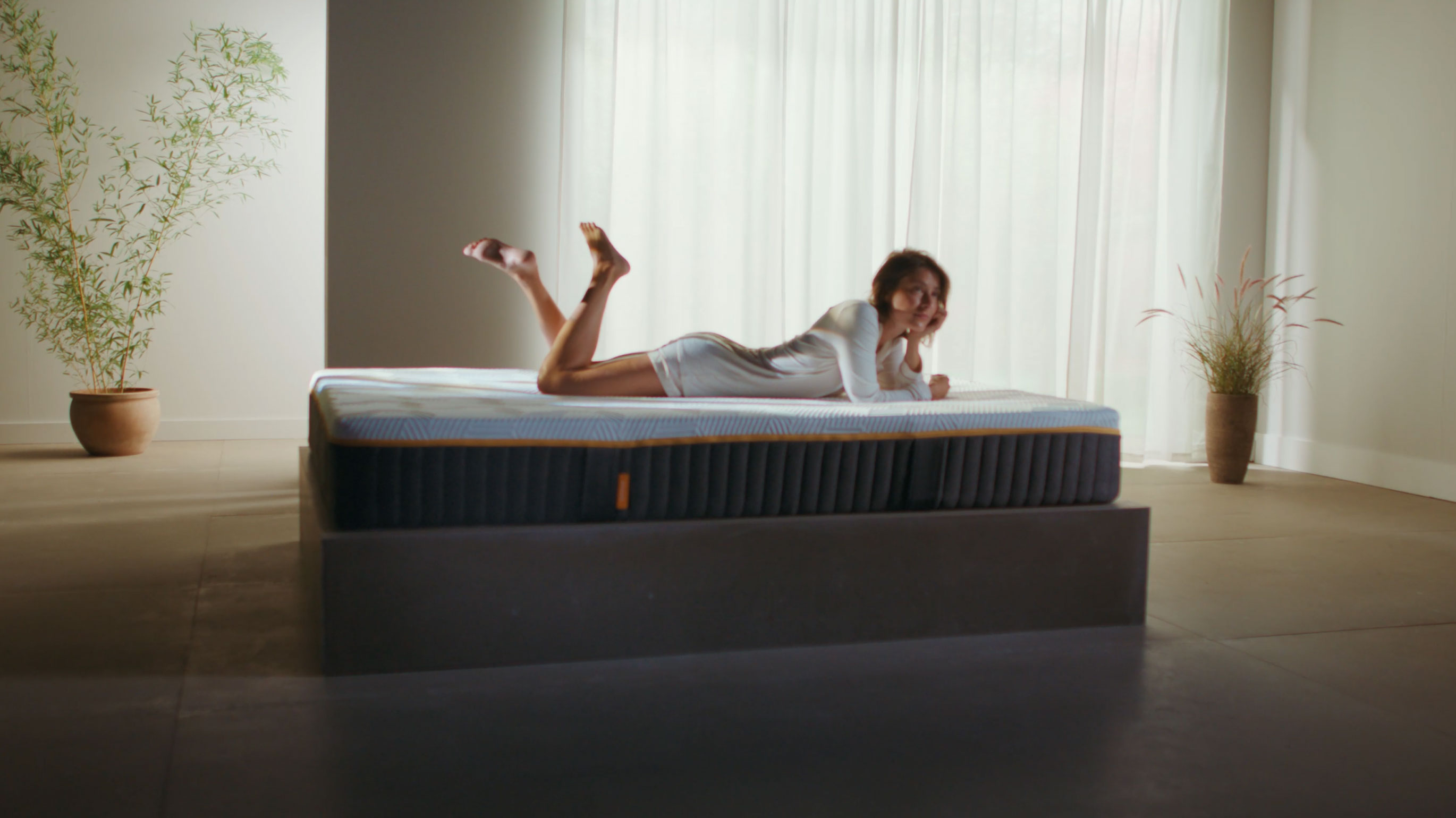 Woman lying on her front on an Emma Original Elite mattress in a bright minimalist room with potted plants.