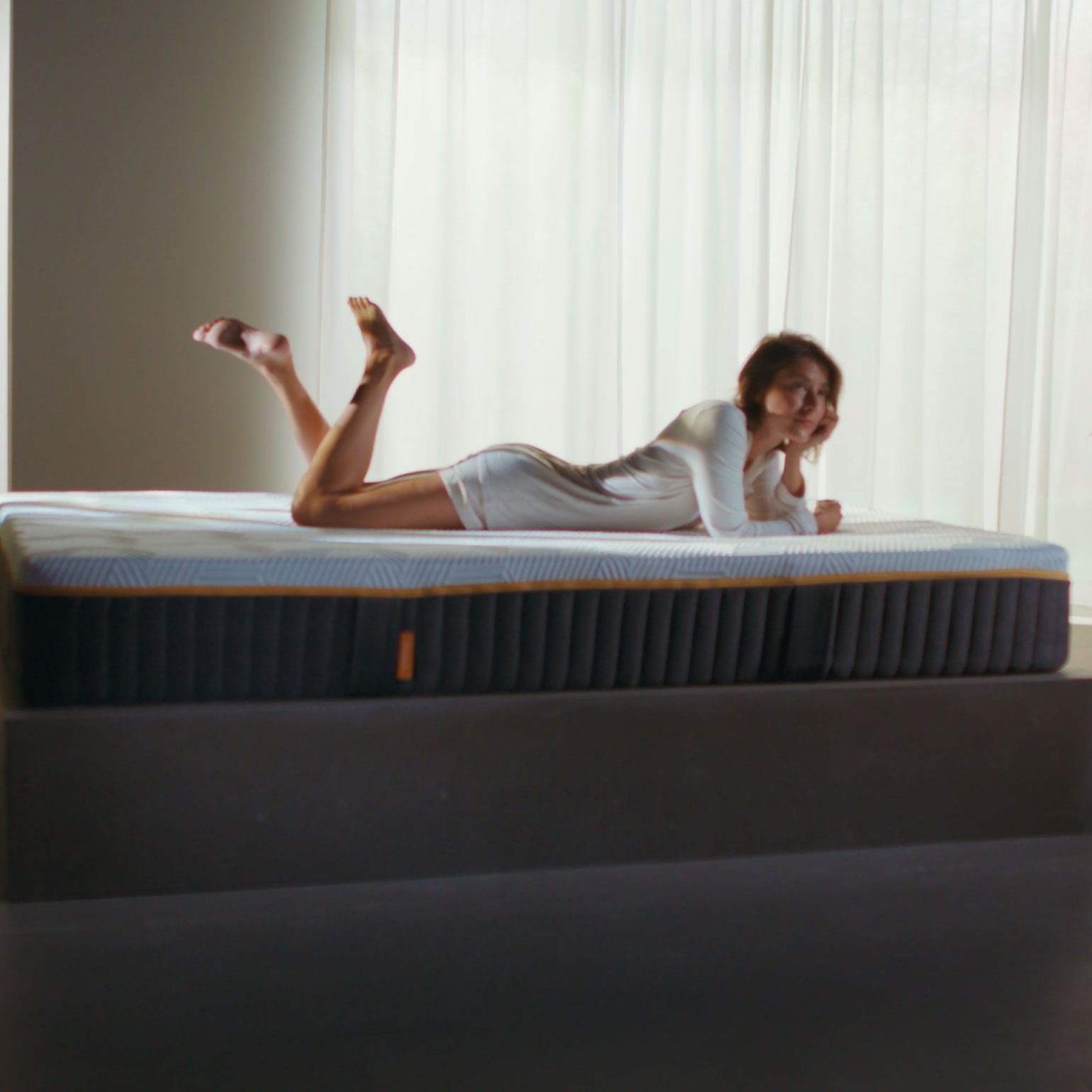 Woman lying on her front on an Emma Original Elite mattress in a bright minimalist room with potted plants.