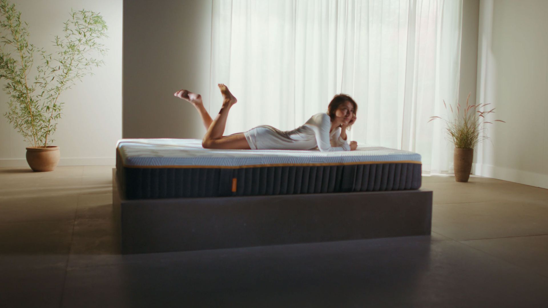 Woman lying on her front on an Emma Original Elite mattress in a bright minimalist room with potted plants.